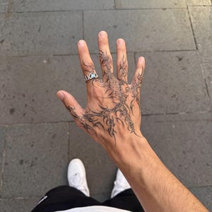 Puede incluir: Un anillo de plata con un diseño texturizado de eslabones abiertos en el dedo anular. La mano presenta un intrincado tatuaje de tinta negra con líneas afiladas, orgánicas y ramificadas que se extienden por el dorso y los dedos.