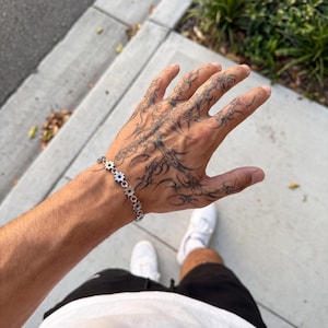 May include: A silver-toned chain bracelet featuring a series of small, interconnected gear-shaped links. The bracelet is worn on a wrist that displays intricate, dark-inked tribal-style tattoo patterns extending across the back of the hand.