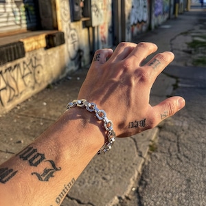 May include: A silver bracelet with a geometric chain design. The bracelet is worn on a person's wrist. The person has tattoos on their arm and fingers. The background is an urban setting with graffiti.