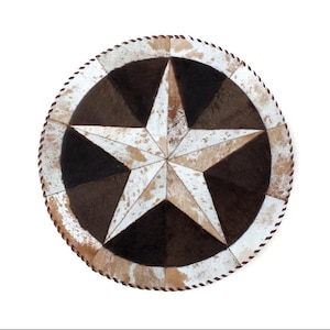 May include: Round cowhide rug featuring a large star design. The star is composed of white, tan, and brown sections, with a dark brown center. The rug's border is a lighter tan with a stitched edge, creating a rustic, Western-inspired aesthetic.