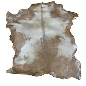 May include: A natural cowhide rug with a light brown base and irregular white patches. The hide has a soft, textured appearance and an organic shape, suitable for home decor.