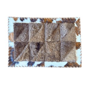 May include: Rectangular rug with a patchwork design. The center features squares of textured, brown material. The border is a mix of white, brown, and black cowhide, with a stitched edge. The rug is on a white background.