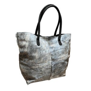 May include: A gray and white cowhide tote bag with black leather handles. The bag has a rectangular shape with a soft, textured surface. The handles are attached to the top of the bag with black leather accents.