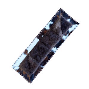 May include: A rectangular, decorative pillow with a brown, shaggy center and a cowhide pattern border. The cowhide features white, brown, and black patches. The edges are trimmed with a stitched, light brown material. The pillow is designed for home decor.