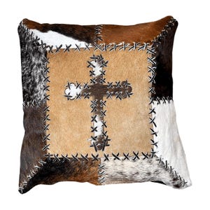 May include: Square decorative pillow with a patchwork design. The pillow features a central tan square with a stitched brown cross. The surrounding patchwork includes brown, black, white, and tan cowhide pieces, all stitched together with black thread.