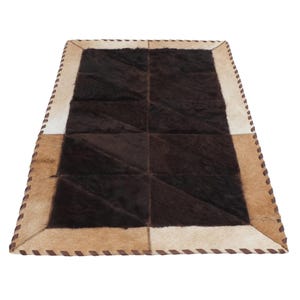May include: Rectangular cowhide rug with a patchwork design. The rug features a dark brown center, surrounded by tan, cream, and brown sections. The edges are stitched with a brown thread, creating a decorative border. The rug is designed for floor use.