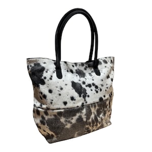 May include: A tote bag with a cowhide pattern in black and white. The bag has black handles and a rectangular shape. The bottom portion of the bag has a darker, more mottled pattern than the top.