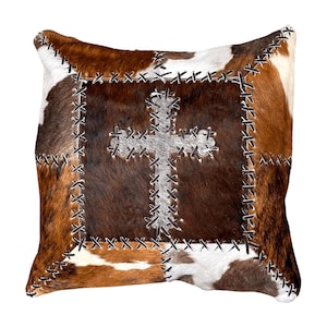May include: Square cowhide pillow with a patchwork design in shades of brown, white, and tan. A silver cross is stitched in the center. The edges are sewn with black thread, creating a rustic, country aesthetic.