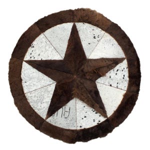 May include: A round rug with a star design. The rug features a large, dark brown star in the center, surrounded by a white and black speckled pattern. The outer edge is a darker brown, creating a distinct border. The rug is made of a cowhide material.