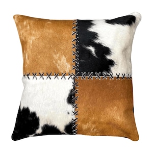 May include: Square decorative pillow with a patchwork design. The pillow features four sections of cowhide in shades of brown, black, and white, stitched together with dark thread. The pillow is square and has a soft, textured appearance.