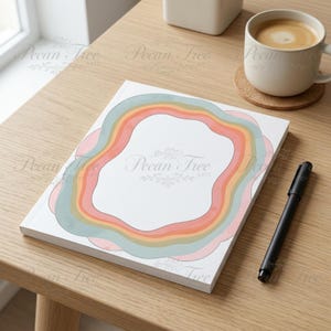 May include: A white notepad with a colorful rainbow design and the text "Pecan Tree Arts" sits on a wooden table. A black pen and a cup of coffee are also on the table. The notepad has a pastel rainbow border.