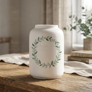 May include: A white ceramic vase with a green leaf wreath design. The vase is cylindrical with a slightly rounded top and sits on a wooden table. The wreath is composed of green leaves and small berries.