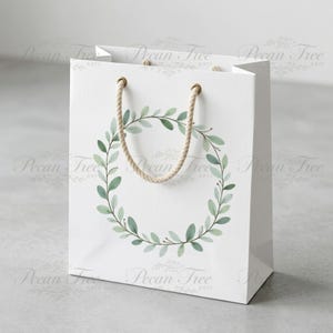 May include: A white gift bag with a watercolor wreath design. The wreath is composed of green leaves and stems. The bag has a rope handle and gold-colored eyelets. The bag is a square shape.