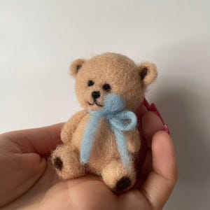 Può includere: Un piccolo orsacchiotto marrone chiaro con occhi di perline nere e una bocca cucita. L'orso ha un nastro azzurro legato al collo. L'orso è fatto di un materiale soffice e ha macchie marroni scure sulle zampe e sulle orecchie.