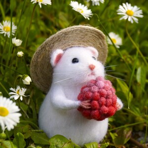 May include: A needle-felted white hamster figurine wearing a small tan woven straw hat. The hamster has pink inner ears and a pink nose, and it is holding a textured, round, bright red raspberry-like object in its small front paws.