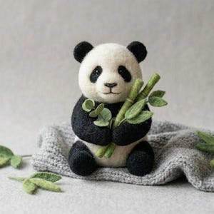 Op de afbeelding: Een naaldgevilte panda-figuur van zwarte en witte wol, met zwarte oren, oogvlekken, armen en benen. De panda houdt verschillende groene gevilte bamboestengels met bladeren vast, met extra losse groene gevilte bladeren in de nabijheid.