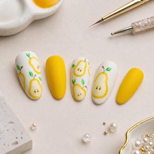May include: A set of five artificial nails. Three nails are white with yellow pear designs and green leaves. Two nails are solid yellow. A small paint palette and nail art tools are in the background.