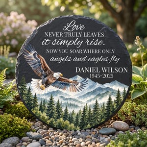 May include: A round black slate memorial plaque featuring a bald eagle in flight over a mountain and forest landscape. The text reads: Love NEVER TRULY LEAVES it simply rise. NOW YOU SOAR WHERE ONLY angels and eagles fly DANIEL WILSON 1945-2025.