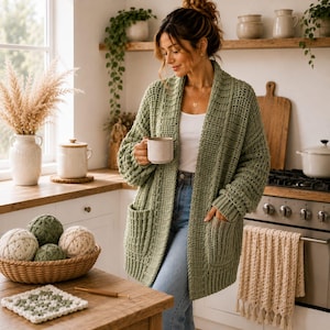 May include: A sage green open-front cardigan with a textured waffle-knit pattern, long sleeves, and two front patch pockets, worn over a white top and blue jeans, while holding a light-colored ceramic mug in a kitchen setting.