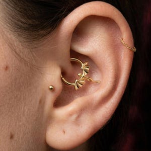 Puede incluir: Primer plano de una oreja con un pendiente dorado en forma de dragón. El pendiente es un diseño de aro con las alas del dragón extendidas. También es visible un pequeño perno dorado. El tono de piel es claro.