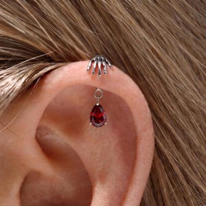 Può includere: Un piercing per orecchio color argento caratterizzato da un design a mano scheletrica nella parte superiore e da un ciondolo a forma di goccia con una gemma rosso scuro, sospeso a un piccolo anello metallico sul cartilagine superiore.