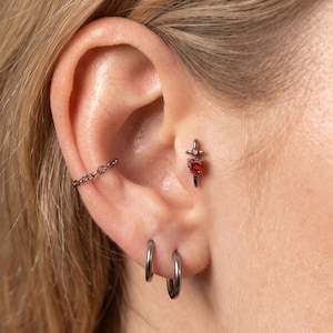 May include: A silver-toned ear cartilage piercing featuring a small red heart-shaped gemstone set within a cross-shaped metal frame. The ear also displays a thin silver chain cuff on the upper cartilage and two plain silver hoop earrings in the lobe.