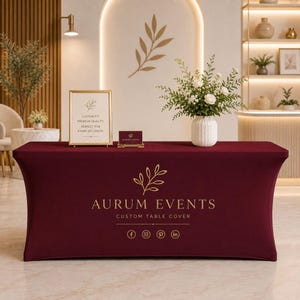 May include: A rectangular burgundy stretch table cover featuring a gold logo with a leaf graphic and the text AURUM EVENTS CUSTOM TABLE COVER, along with social media icons. The fabric fits tightly over the table, creating a smooth, clean surface.