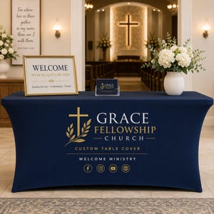 May include: A navy blue fitted table cover featuring the white and gold Grace Fellowship Church logo, text, and social media icons. The table is set with a framed welcome sign, a small card holder, and a white vase filled with white flowers.