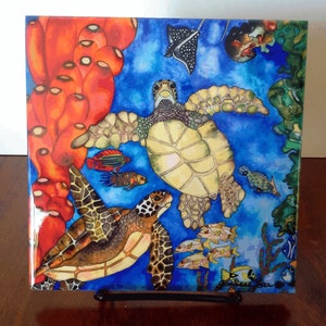 May include: A colorful ceramic tile with a watercolor painting of two sea turtles swimming in a blue ocean. The tile features coral, fish, and a stingray.