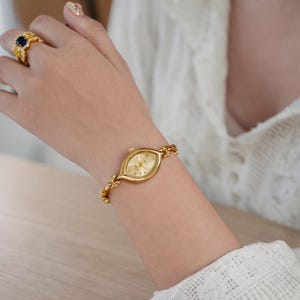 May include: A gold-toned wristwatch with an oval face and a delicate chain-link band. The watch face has gold hands and markers. The band features floral accents. A gold ring with a blue stone is also visible.