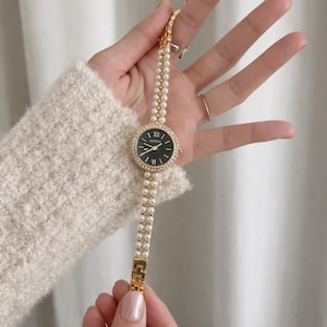 May include: A gold-toned wristwatch with a black face and a bezel encrusted with small, clear crystals. The watch band is composed of two strands of white pearls. The watch is held by a person wearing a cream-colored sweater.
