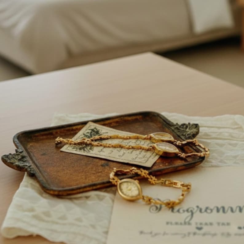 May include: A vintage-style tray with ornate edges holds two gold-toned wristwatches and a gold chain necklace. The watches have oval faces and are displayed on a cream-colored lace cloth and a vintage card.