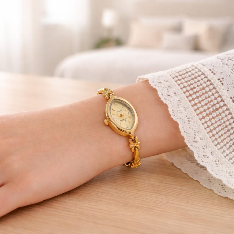 May include: A gold-toned wristwatch with an oval face and a delicate floral design on the band. The watch has a light-colored face and is worn on a wrist. The band is approximately 6 inches in length.