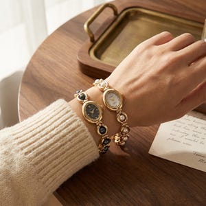 May include: Two gold wristwatches with oval faces and pearl and black bead detailing. One watch has a black face, the other a white face. The watches are on a wrist, with a wooden tray and a cream-coloured sweater in the background.