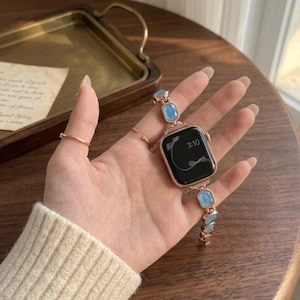 Op de afbeelding: Een roségouden smartwatchband met rechthoekige lichtblauwe stenen schakels. De wijzerplaat is zwart met een digitaal display dat de tijd 3:10 en een witte lijngrafiek toont. De band is verbonden met een metalen roségouden horlogekast.