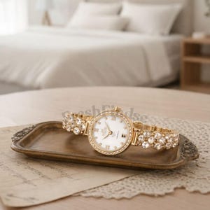 Puede incluir: Un reloj de pulsera dorado con esfera blanca, números romanos y bisel y correa con incrustaciones de cristal. El reloj se exhibe en una bandeja decorativa con un tapete de encaje y un trozo de papel.