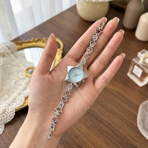 May include: A silver-toned analogue wristwatch featuring a star-shaped case with a light blue dial. The watch band is constructed from a series of interconnected silver metal links, alternating between open star and heart-shaped outlines throughout.