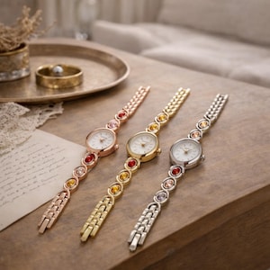 May include: Three elegant women's watches with round faces and decorative bands. The watches are in rose gold, gold, and silver tones, each adorned with colourful gemstones. The watches are displayed on a wooden surface.