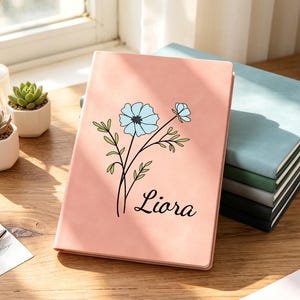 Puede incluir: Un diario rosa con un diseño floral y el nombre "Liora" en cursiva. El diario está abierto, revelando una pila de otros diarios en tonos verdes, grises y negros. También se ven suculentas en macetas blancas y un bolígrafo.