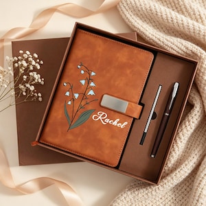 May include: A brown leather journal and pen set in a gift box. The journal features a floral design and the name "Rachel" in white script. The set includes a silver pen and a dark brown pen.