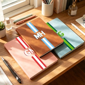 May include: Three personalised notebooks with initials and stripes in red, blue, and green. The notebooks are on a wooden surface, with pens, a succulent plant, and other stationery items nearby. The notebooks are in shades of pink, brown, and blue.