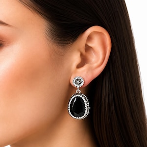 Peut inclure: Une boucle d'oreille pendante composée d'un clou circulaire et d'un grand pendentif en forme de poire noire. La monture en métal argenté est incrustée de petits cristaux clairs et étincelants qui bordent le clou circulaire et la pierre noire.
