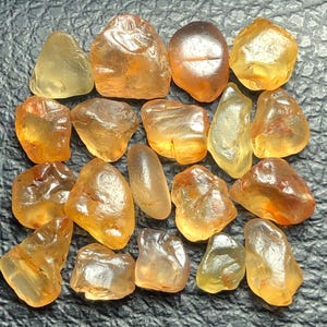 Op de afbeelding: Een verzameling van zeventien losse, getrommelde edelstenen in diverse tinten oranje, geel en amber. Elke steen heeft een glad, gepolijst oppervlak met natuurlijke variaties in helderheid en kleur, variërend van licht doorschijnend geel tot dieporanje.