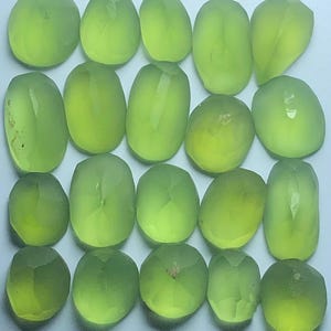 May include: A collection of twenty translucent, light green gemstones arranged in a four-by-five grid. Each stone features a faceted surface with a smooth, rounded, oval or pear-like shape, reflecting light across their polished, pale lime-coloured surfaces.