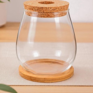 Peut inclure: Un bocal en verre transparent avec un couvercle en liège, posé sur une base ronde en bois. Le bocal a une forme large et arrondie, et le couvercle en liège a une ouverture circulaire au centre.