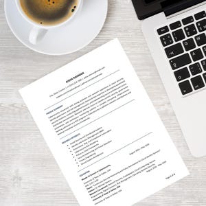 May include: A resume titled "AISHA RAHMAN" is displayed on a wooden desk next to a laptop and a cup of coffee. The resume lists professional experience and education. The laptop is open, and the coffee cup is white.