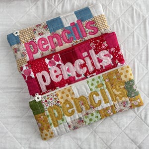 May include: Three patchwork pencil cases, each embroidered with the word "pencils". The cases feature a variety of fabrics in shades of blue, pink, red, yellow, and green, with floral and polka dot patterns. Each case has a zip closure.