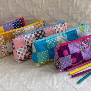 May include: A selection of patchwork pencil cases in various colours, each with a zip closure. The cases are made from patterned fabrics in pink, yellow, blue, and purple. Pencils and pens are arranged in front of the cases.
