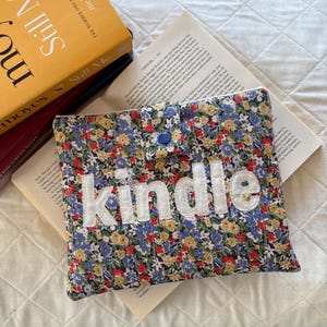 May include: A quilted fabric sleeve featuring a vibrant floral pattern with blue, yellow, and red flowers. The sleeve has a flap closure secured with a blue snap button and the word kindle is appliqued in white fabric letters across the front.