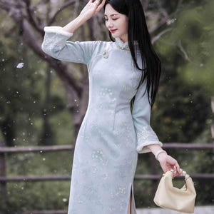 May include: A light blue, long-sleeved dress with a floral pattern and a high collar. The dress features white fur trim on the cuffs and a side slit. A small, cream-colored handbag with pearl handles is held in the hand.